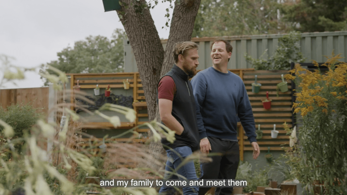 two people walk through a garden, one smiling and one looks serious