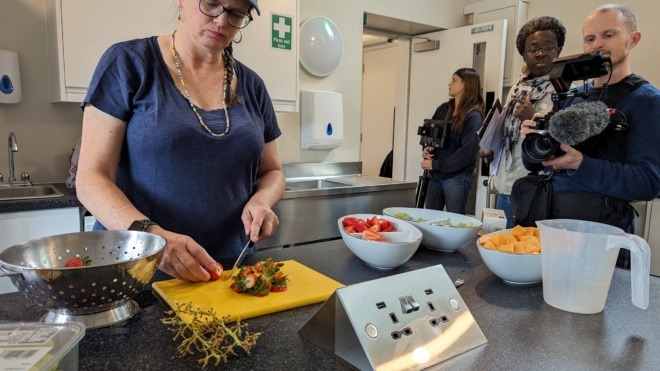 woman in kitchen preparing food, man holding camera filming