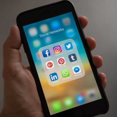 A hand holding an iPhone showing a number of social media apps