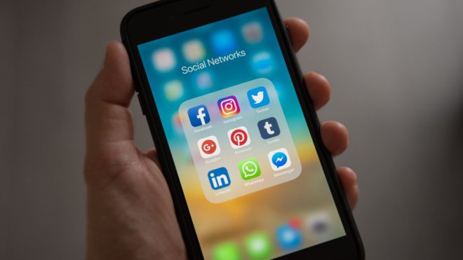 A hand holding an iPhone showing a number of social media apps